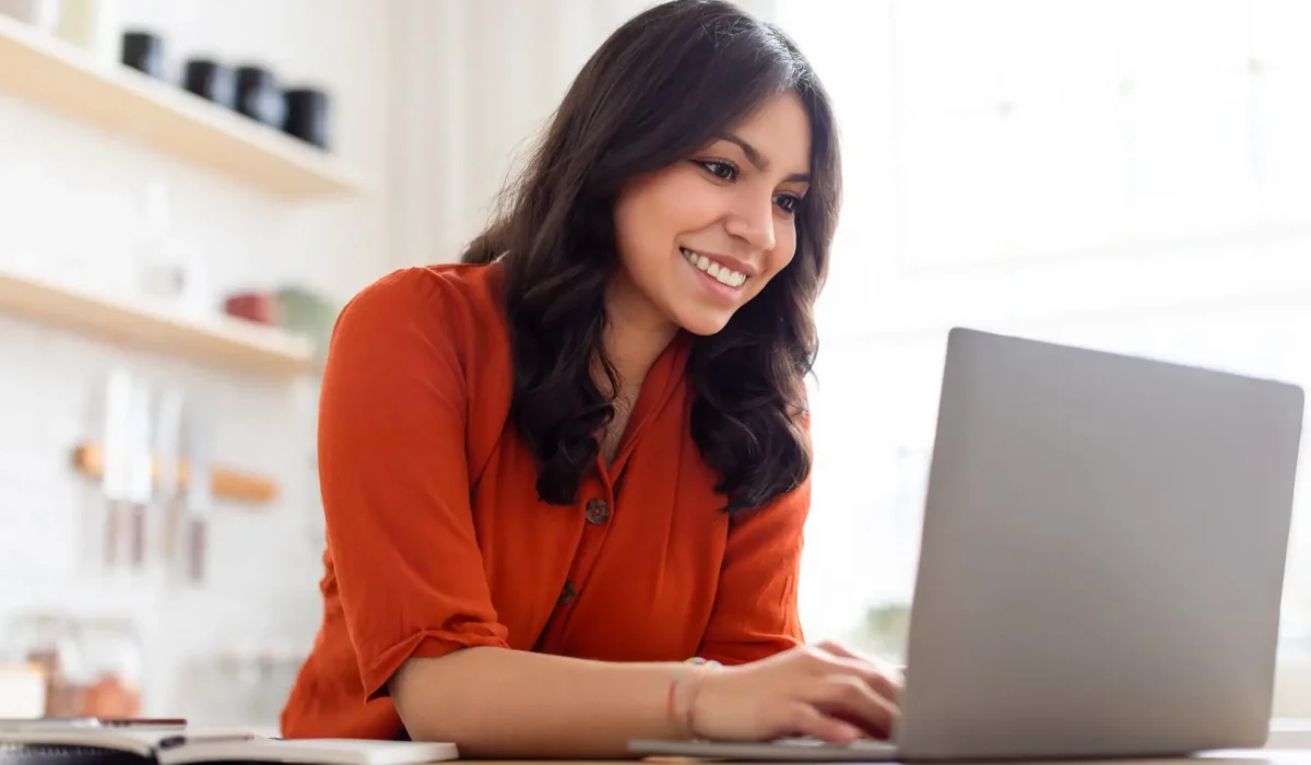 Joyful middle eastern woman with brown hair working on a laptop and smiling in a home environment at kicthen